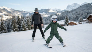 Enfant souriant en combinaison de ski descendant une piste débutant en chasse-neige, un parent skiant derrière lui