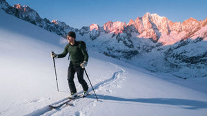 Skieur de randonnée en première couche en laine mérinos verte montant en peaux de phoque sur une pente enneigée au lever du soleil, sommets roses en arrière-plan