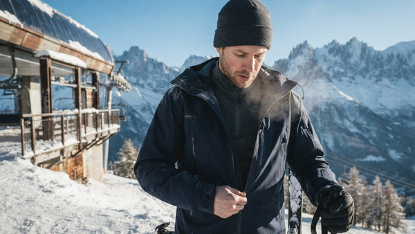 Skieur au sommet d'un télésiège fermant sa veste de ski par-dessus une première couche en laine mérinos à col zippé, par un matin d'hiver ensoleillé
